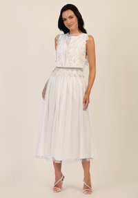Vestido blanco de encaje con mangas rizadas, detalles intrincados, falda de tul con puntos y una cintura ajustada. Combinado con tacones plateados de tiras.