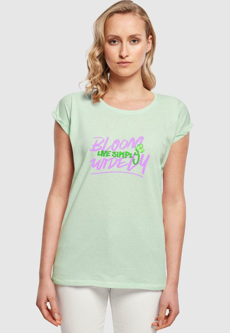 Mintgrøn kortærmet t-shirt med opkrøllede ærmer, prydet med et grafisk print i pink og grøn, der siger "BLOOM LIVE SIMPLY WIDELY."