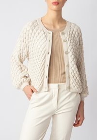 Teksturirani krem kardigan s dijamantnim uzorkom, okruglim ovratnikom i zatvaranjem na gumbi, uparen s bež rebrastim topom i bijelim hlačama.