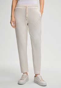 Light beige jogger pants with an elastic waistband and drawstring, featuring side pockets and a subtle logo embroidery. Smooth texture.