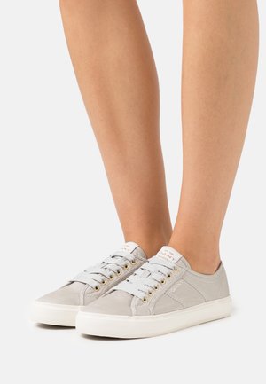 Vaaleanharmaat kangaskengät valkoisilla kumipohjilla, joissa on pyöreät kultaiset reiät, litteät nauhat ja logolla varustettu labeli kielekkeessä.