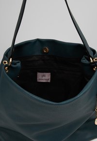 Tyrkysová kožená kabelka s hladkou texturou, černým vnitřkem, zlatými kovovými detaily a značkovou štítkem. Otevřený design a popruh.