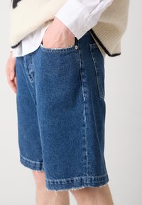 Shorts en denim bleu avec un ourlet effiloché, dotés de poches latérales et d'une coupe décontractée, associés à une chemise blanche et à un pull en tricot.