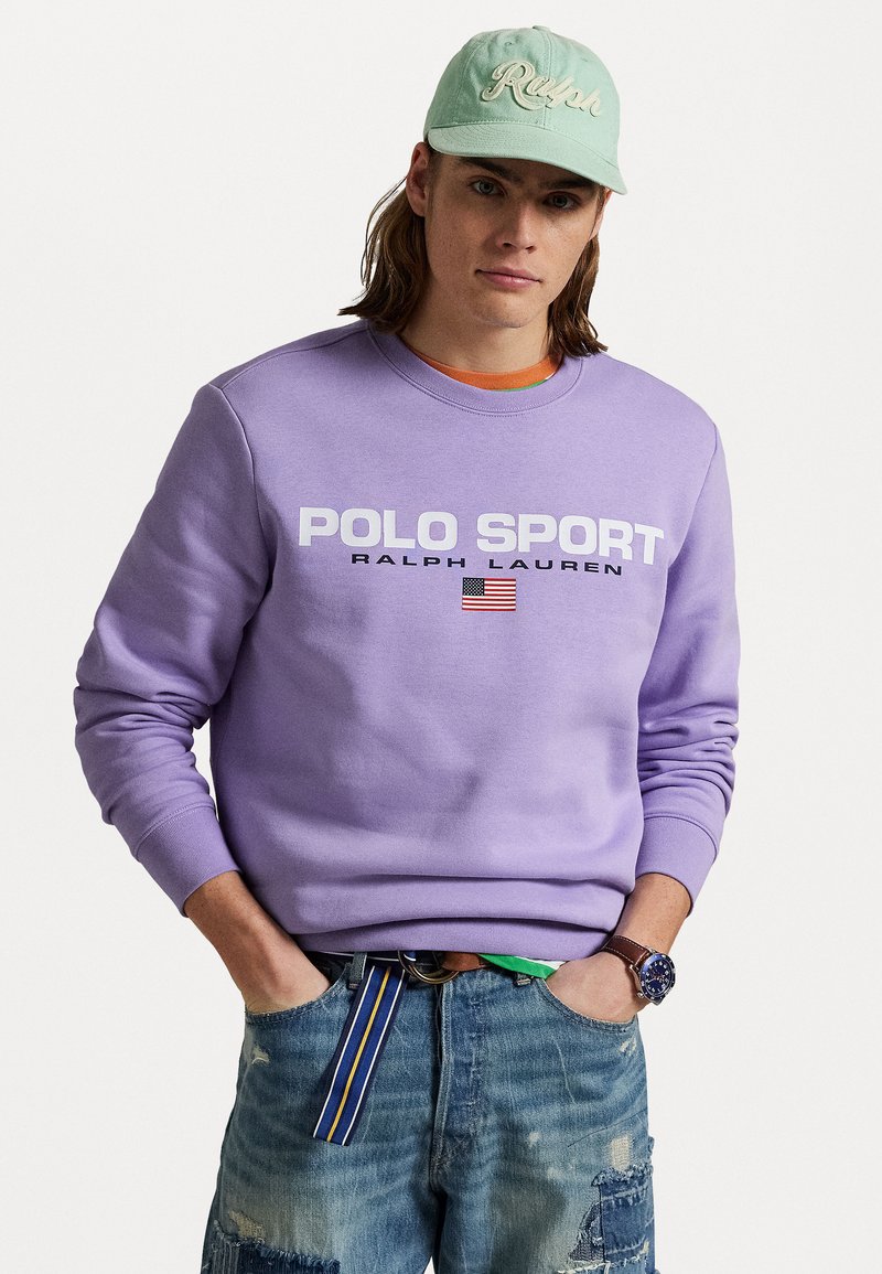 Fialová mikina s logom "POLO SPORT", prednou grafikou a americkou vlajkou. Modré roztrhnuté džínsy a mintová zelená šiltovka.