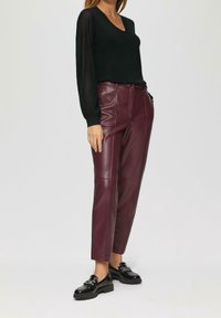 Kobieta ubrana w czarną bluzkę z dekoltem w serek i przezroczystymi długimi rękawami, bordowe skórzane spodnie oraz czarne lakierowane loafersy, stojąca na białym tle.