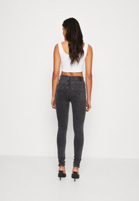 Mujer con cabello largo y ondulado, vistiendo una blusa sin mangas blanca de corte corto, jeans skinny de cintura alta negros y zapatos de tacón alto negros, mirando hacia atrás.