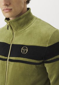 Zelena velur zip-jakna s crnim horizontalnim prugama i zlatnim izvezenim logotipom na prsima. Ima visoki ovratnik i mekanu teksturu.