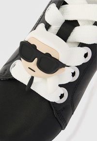 Μαύρο αθλητικό παπούτσι με άσπρα κορδόνια, με ένα πλαστικό γούστο που απεικονίζει μια φιγούρα με λευκά μαλλιά και γυαλιά ηλίου στα κορδόνια.