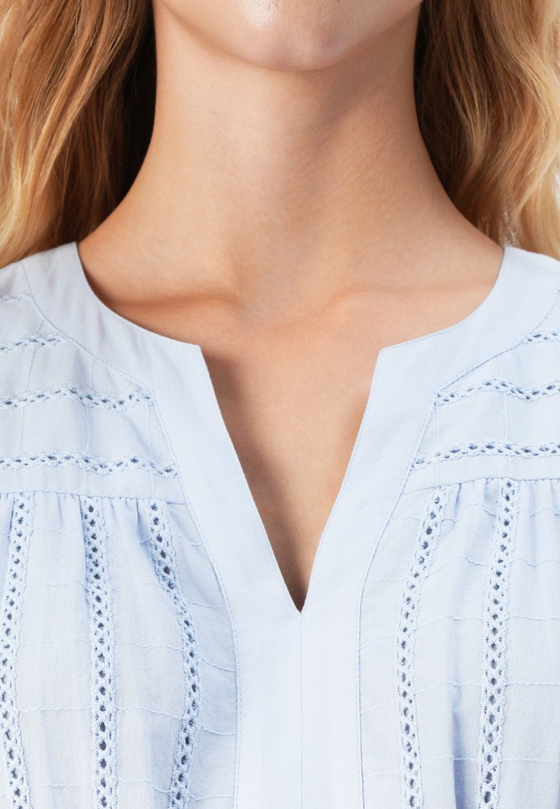 Zbliżenie na szyję i górną część klatki piersiowej kobiety noszącej jasnoniebieską bluzkę z pionowymi haftowanymi koronkami i dekoltem w kształcie litery V.