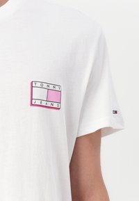 T-shirt en coton blanc avec un petit logo rectangulaire en rose, noir et blanc sur la poitrine gauche. La manche présente un accent rayé multicolore.