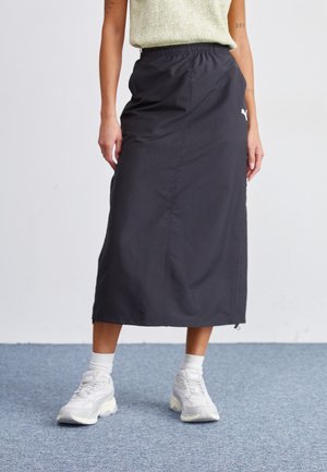 Personne portant une jupe noire mi-longue avec une ceinture élastique, des baskets blanches, des chaussettes blanches et un haut à manches courtes vert clair.