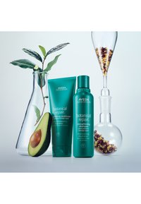 Aveda botanisk reparerande stärkande schampo och balsam i turkosa tuber, bredvid en avokado och en glasbehållare med frön.