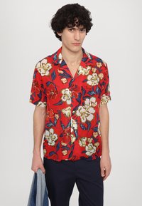 Camisa roja de manga corta con un patrón floral de grandes flores en color crema y azul marino, diseño con cuello y cierre de botones.