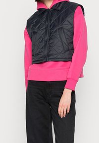 Schwarze gesteppte Weste mit Kragen, kombiniert mit einem leuchtend pinken Hoodie und schwarzen Jeans. Verfügt über Druckknopfverschlüsse und zwei Vorder pockets.