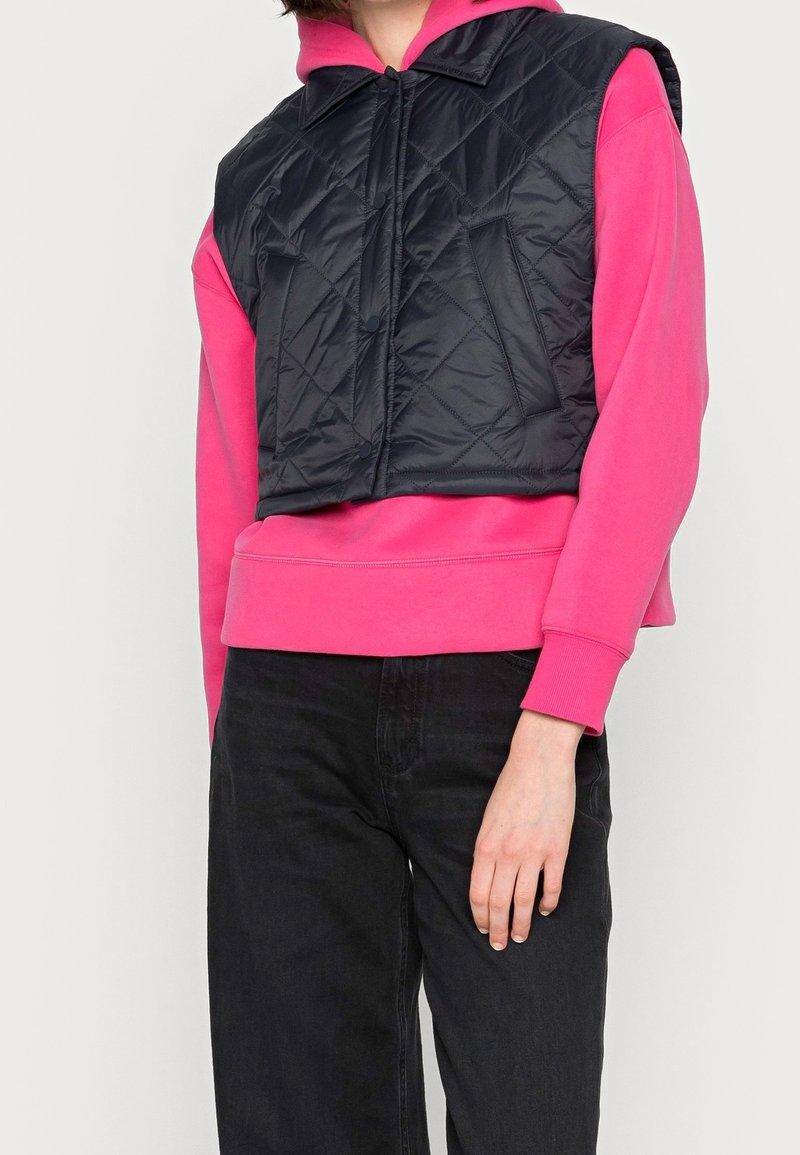 Schwarze gesteppte Weste mit Kragen, kombiniert mit einem leuchtend pinken Hoodie und schwarzen Jeans. Verfügt über Druckknopfverschlüsse und zwei Vorder pockets.