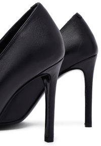 Zapatos de tacón alto de cuero negros con punta afilada y tacones delgados tipo stiletto vistos desde la parte trasera sobre un fondo blanco.
