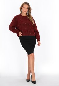 Pullover tricotat de culoare burgundă, cu mărgele decorative albe, combinat cu o fustă neagră ajustată. Modelul poartă tocuri negre înalte. Textură netedă.