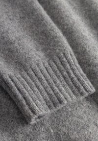 Grauer Wollpullover mit einem gerippten Bund, der eine weiche Textur und eine leicht flauschige Oberfläche zeigt. Der Stoff wirkt warm und gemütlich.