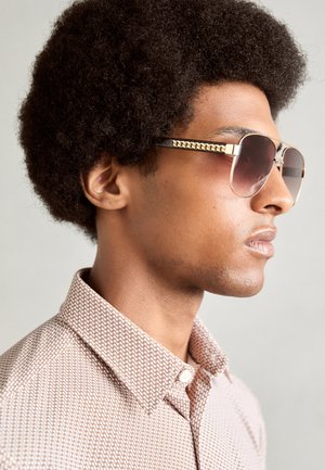 Sunglasses with brown gradient lenses and gold hardware, featuring chain-like detail along the temples. Model wears a patterned shirt.