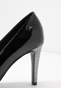Zapato de tacón alto de charol negro con un tacón geométrico texturizado en plata y un pequeño logo embosado en el costado.