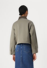 Giacca corta verde oliva con colletto marrone, vestibilità rilassata e polsini con bottoni, abbinata a una gonna di jeans blu con tasche.