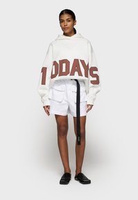 Haut à capuche oversized blanc avec des lettres en bloc marron "100 JOURS", assorti à un short cargo blanc et des chaussures noires à enfiler. Tissu lisse, coupe décontractée.
