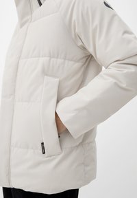 Veste rembourrée beige clair avec une main dans la poche, une fermeture éclair noire près de l'épaule et une petite étiquette noire sur le bord de la poche.