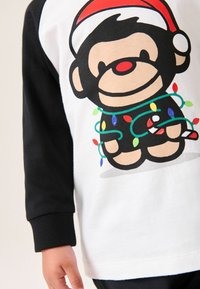 Weißes T-Shirt mit schwarzen Ärmeln, das einen Cartoon-Affen in einem Santa-Hut zeigt, der in bunten Weihnachtslichtern eingewickelt ist und einen Zuckerstangen hält.