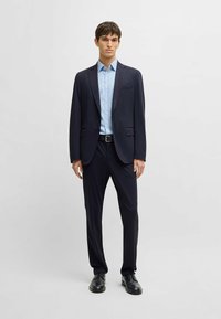 Marineblauwe suit, bestaande uit een aansluitende blazer en broek, gecombineerd met een lichtblauw overhemd met knopen. Glad materiaal, klassieke vorm en minimalistisch ontwerp.