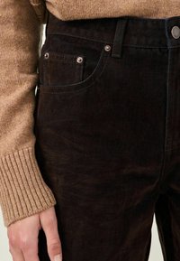 Tmavě hnědé manšestrové kalhoty s pěti kapsami, detaily z kovových nýtů a viditelným texturálním kontrastem oproti světle hnědému pletenému svetru.