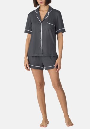 Kobieta stoi boso, ubrana w ciemnoszary krótki, zapinany na guziki top od piżamy i dopasowane szorty z białą lamówką na krawędziach i kieszeni.