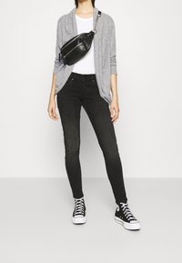 Szary kardigan, biała koszulka, czarne obcisłe dżinsy i czarne sneakersy. Torebka noszona na ramieniu z srebrnymi okuciami. Gładkie tekstury i neutralne kolory.