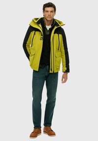 Gul og sort vandtæt jakke med flere lommer, lynlåslukninger, justerbar hætte, lagdelt over en sort skjorte, kombineret med blå jeans og brune sko.