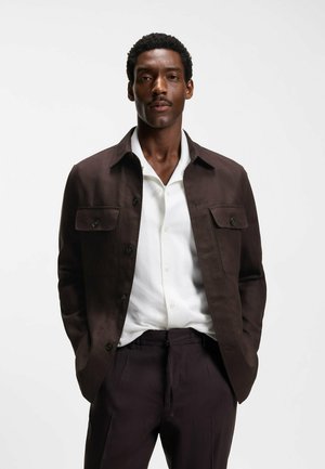 Homme portant une veste marron foncé sur une chemise blanche boutonnée et un pantalon marron foncé assorti, debout les mains dans les poches devant un fond uni.