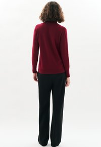 Persoon met krullend haar, gekleed in een rode lange mouwen sweater en zwarte wijde broek, staand met de rug naar de camera tegen een effen witte achtergrond.