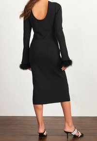 Abito in maglia nera con maniche lunghe, caratterizzato da polsini fluffy, scollatura a forma di barca sul retro e silhouette aderente, che arriva a metà polpaccio.