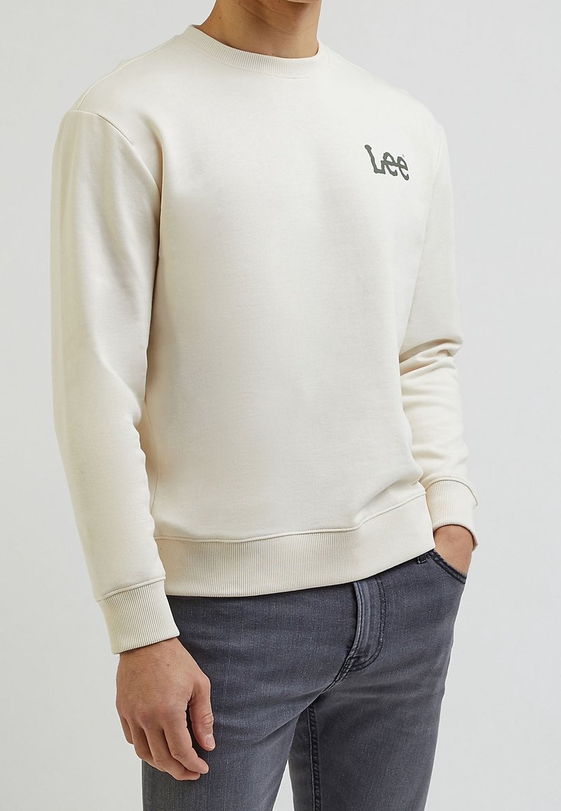 Homme portant un sweat-shirt Lee à manches longues de couleur blanc cassé et un jean gris foncé, debout avec une main dans la poche, devant un fond uni.
