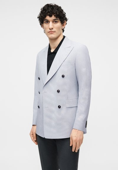 Νεαρός με σγουρά μαλλιά που φοράει ένα ανοιχτό γκρι σακάκι διπλής σειράς κουμπιών πάνω από μαύρο πουκάμισο, στέκεται μπροστά σε απλό φόντο.