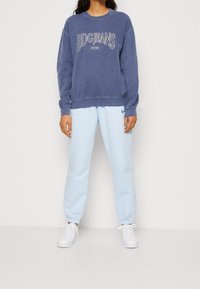 Person som bär en blå BDG Jeans sweatshirt, ljusblå Nike träningsbyxor och vita sneakers medan de står mot en helt vit bakgrund.
