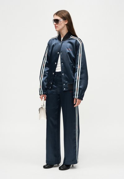 Γυναίκα που φοράει σκούρο μπλε σατινέ μπουφάν και παντελόνι με λευκές πλαϊνές ρίγες, μαύρες μπότες, μεγάλα γυαλιά ηλίου και κρατά ένα μικρό λευκό τσαντάκι.