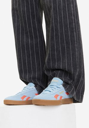 Svetlomodré semišové tenisky s červenými detailmi a podrážkou z gumovej gumy. Nosené s širokými nohavicami so sivými pruhmi.
