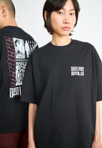 Black cotton t-shirt with oversized fit. Front features small white logo; back has graphic design with red and white elements. Short sleeves.