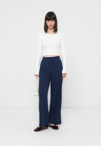 Witte lange mouwen crop top gecombineerd met marineblauwe wijde broek, aangevuld met een kleine witte handtas en glanzende bordeauxrode flats.