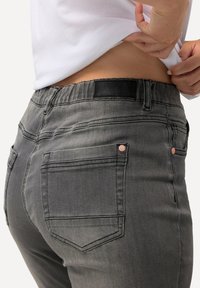 Vaqueros de mezclilla gris elásticos con una cintura alta, dos bolsillos traseros y detalle del logo en la cintura, mostrando una textura suave.