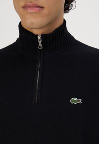 Pulover tricotat de culoare albastru închis, cu guler înalt cu model împletit, închidere cu fermoar pe jumătate și un mic logo verde de crocodil pe partea stângă a pieptului.