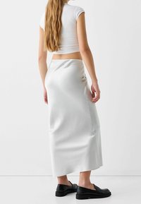 Bílá saténová maxi sukně s hladkým povrchem, se bočním rozparkem, v kombinaci s černými mokasínami. Na sobě má fitted bílý crop top.