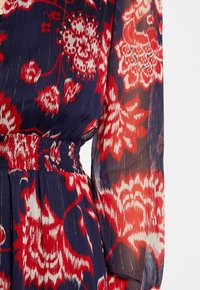 Robe florale avec un fond bleu marine, ornée de motifs de fleurs rouges et blanches, texture plissée et taille élastiquée avec des manches longues.