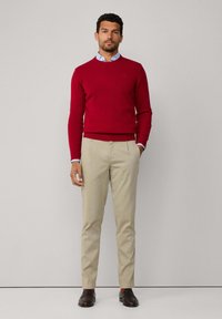 Homme debout contre un mur clair, portant un pull rouge par-dessus une chemise bleu clair, un pantalon beige et des chaussures brun foncé avec des chaussettes orange.