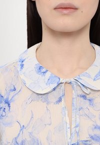 Kwiatowa bluzka wykonana z jasnobeżowego materiału ozdobionego niebieskimi nadrukami w kwiaty. Posiada okrągły kołnierzyk oraz zapięcie na wiązanie przy szyi.