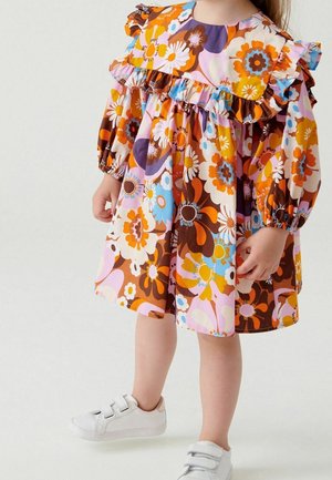 Enfant portant une robe à manches longues avec des motifs floraux orange, marron, rose, bleu et blanc, et des baskets blanches à velcro sur un fond uni.